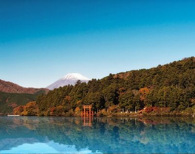 Serene blue lake reflects autumn forests, a red torii gate, and snow-capped Mount Fuji beneath a clear sky in Japan on a calm morning.
