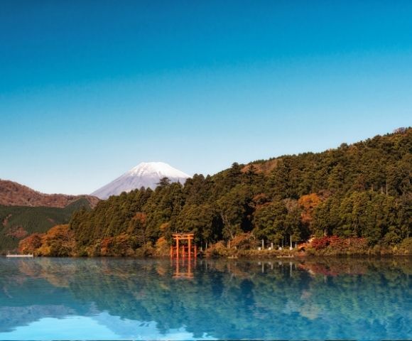 Serene blue lake reflects autumn forests, a red torii gate, and snow-capped Mount Fuji beneath a clear sky in Japan on a calm morning.