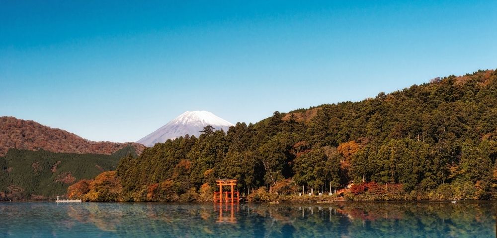 Serene blue lake reflects autumn forests, a red torii gate, and snow-capped Mount Fuji beneath a clear sky in Japan on a calm morning.