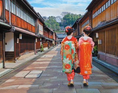 Two women in colorful kimonos walk through a traditional Japanese village street, framed by wooden houses, greenery, and distant hills.