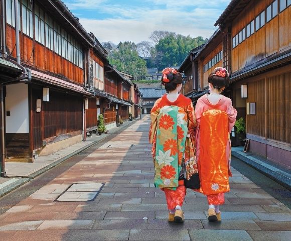 Two women in colorful kimonos walk through a traditional Japanese village street, framed by wooden houses, greenery, and distant hills.