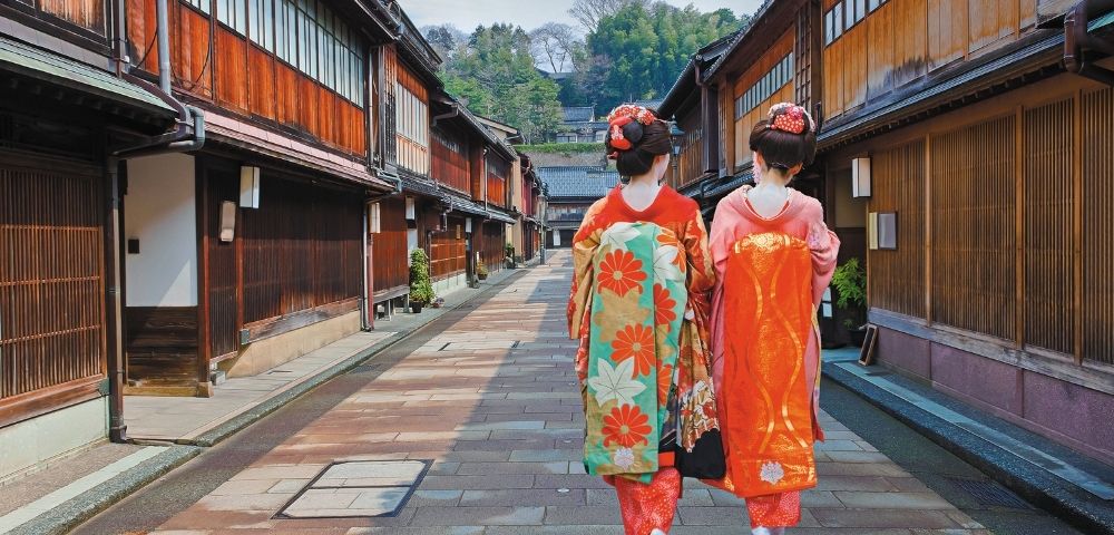 Two women in colorful kimonos walk through a traditional Japanese village street, framed by wooden houses, greenery, and distant hills.