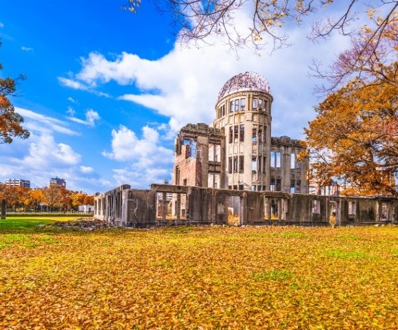 Historic Hiroshima Peace Memorial ruins with a domed tower stand in an autumn park, surrounded by golden leaves under a vivid blue sky.