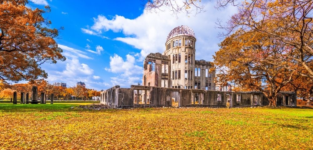 Historic Hiroshima Peace Memorial ruins with a domed tower stand in an autumn park, surrounded by golden leaves under a vivid blue sky.