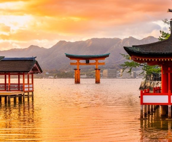 Sunset over Japan’s Itsukushima Shrine, with a red torii gate rising from calm water, shrine buildings, mountains, and glowing orange skies.
