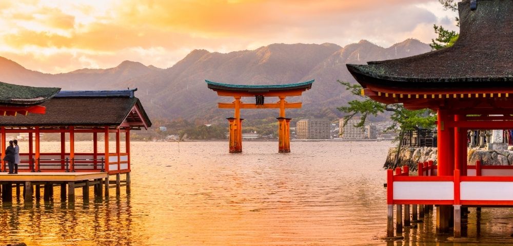 Sunset over Japan’s Itsukushima Shrine, with a red torii gate rising from calm water, shrine buildings, mountains, and glowing orange skies.
