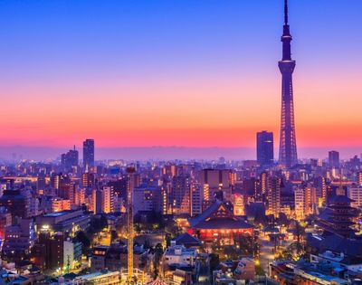 Tokyo skyline at sunset, with Tokyo Skytree towering over glowing city buildings, temples, and streets beneath a vivid pink-blue sky.