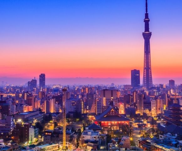 Tokyo skyline at sunset, with Tokyo Skytree towering over glowing city buildings, temples, and streets beneath a vivid pink-blue sky.