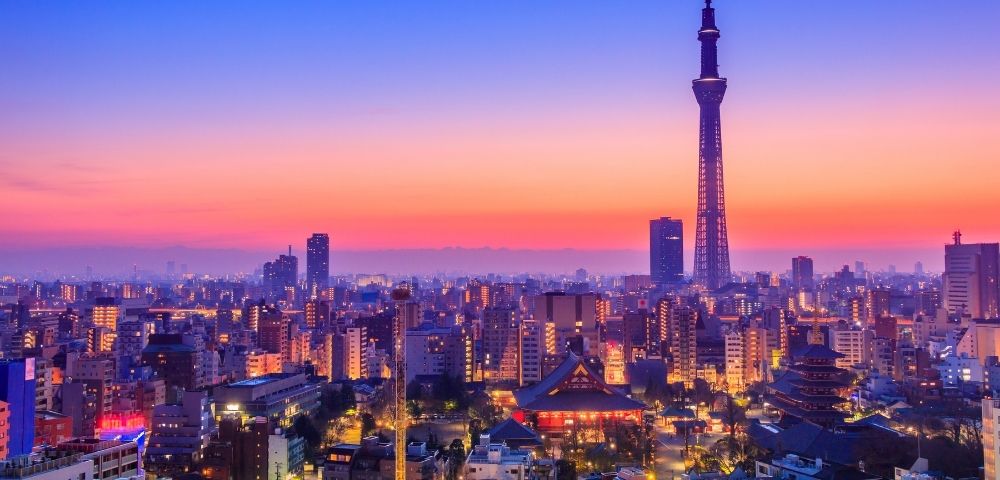 Tokyo skyline at sunset, with Tokyo Skytree towering over glowing city buildings, temples, and streets beneath a vivid pink-blue sky.
