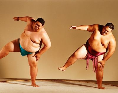Two plus-size male dancers perform energetic balancing poses in colorful loincloths, arms extended and one leg raised, conveying strength and joy.