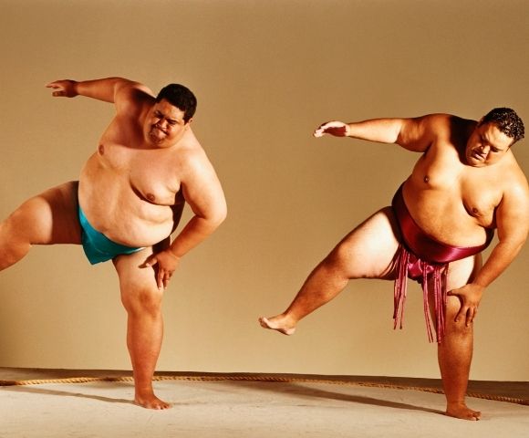 Two plus-size male dancers perform energetic balancing poses in colorful loincloths, arms extended and one leg raised, conveying strength and joy.