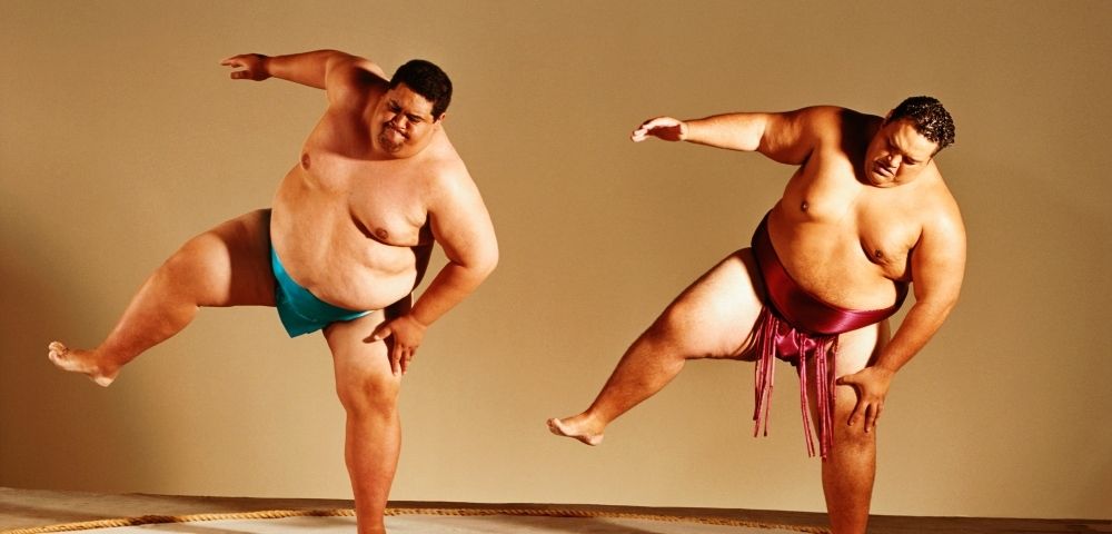 Two plus-size male dancers perform energetic balancing poses in colorful loincloths, arms extended and one leg raised, conveying strength and joy.