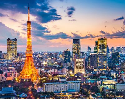 Tokyo Tower glows orange over a vibrant city skyline at sunset, with illuminated skyscrapers, dense buildings, and colorful clouds creating a lively mood.