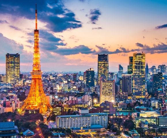 Tokyo Tower glows orange over a vibrant city skyline at sunset, with illuminated skyscrapers, dense buildings, and colorful clouds creating a lively mood.