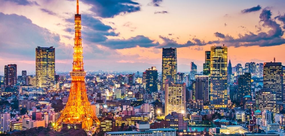Tokyo Tower glows orange over a vibrant city skyline at sunset, with illuminated skyscrapers, dense buildings, and colorful clouds creating a lively mood.