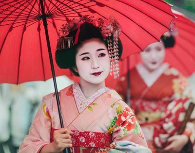 Geisha in a colorful kimono holds a red parasol, with ornate hairpins and pale makeup; another woman in red stands behind her.