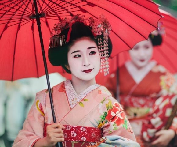 Geisha in a colorful kimono holds a red parasol, with ornate hairpins and pale makeup; another woman in red stands behind her.