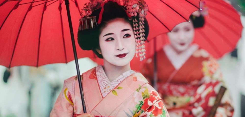 Geisha in a colorful kimono holds a red parasol, with ornate hairpins and pale makeup; another woman in red stands behind her.
