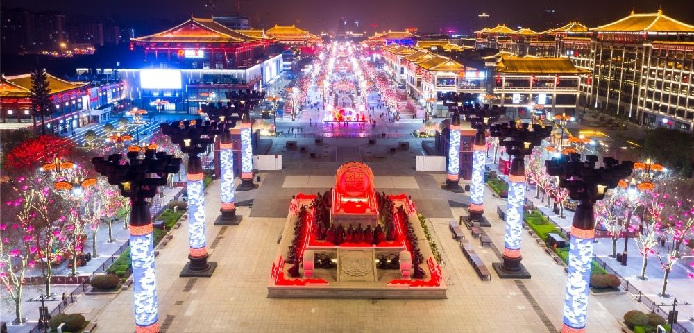 An illuminated street scene at night with traditional Asian architecture, ornate lampposts, and a central monument featuring a large red emblem surrounded by figures.