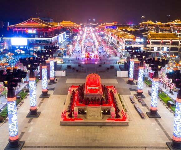 An illuminated street scene at night with traditional Asian architecture, ornate lampposts, and a central monument featuring a large red emblem surrounded by figures.