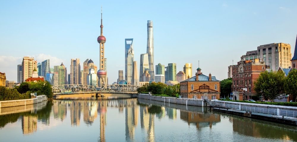 Reflections of a modern city skyline, including a distinctive spherical tower and various skyscrapers, are visible in a calm river in front of an old brick building under a clear blue sky.