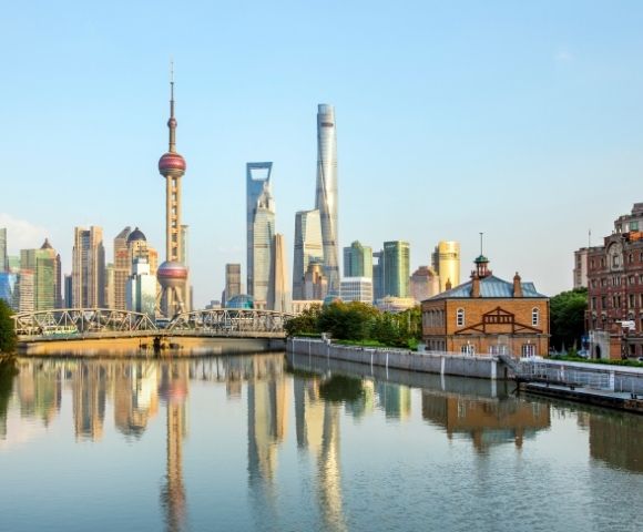 Reflections of a modern city skyline, including a distinctive spherical tower and various skyscrapers, are visible in a calm river in front of an old brick building under a clear blue sky.
