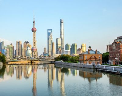 Reflections of a modern city skyline, including a distinctive spherical tower and various skyscrapers, are visible in a calm river in front of an old brick building under a clear blue sky.
