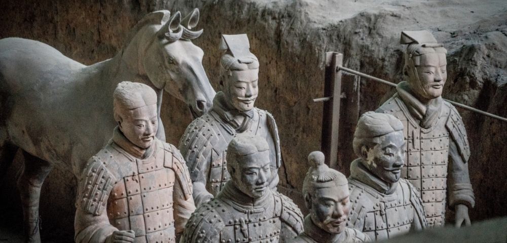 A group of terracotta warrior statues with a horse statue in the background stand in rough, excavated earth.