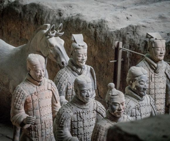 A group of terracotta warrior statues with a horse statue in the background stand in rough, excavated earth.