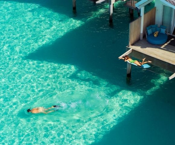 Aerial view of a swimmer in bright turquoise water beside an overwater bungalow and shaded dock, conveying a peaceful tropical escape.