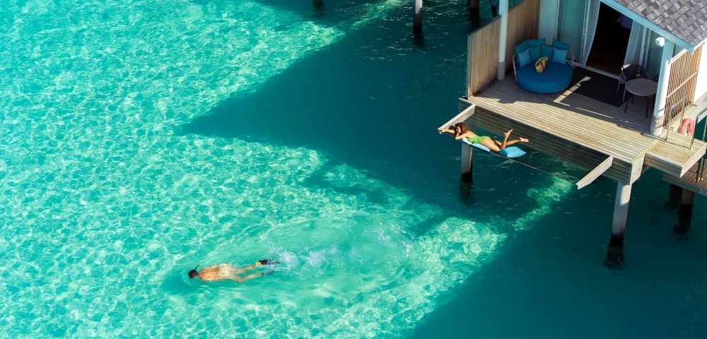 Aerial view of a swimmer in bright turquoise water beside an overwater bungalow and shaded dock, conveying a peaceful tropical escape.