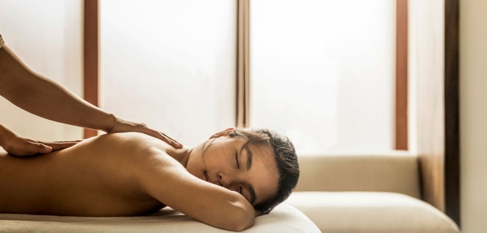 Shirtless man rests face down on a sofa while receiving a back massage in a softly lit room, creating a calm, relaxing atmosphere.