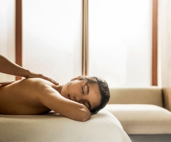 Shirtless man rests face down on a sofa while receiving a back massage in a softly lit room, creating a calm, relaxing atmosphere.