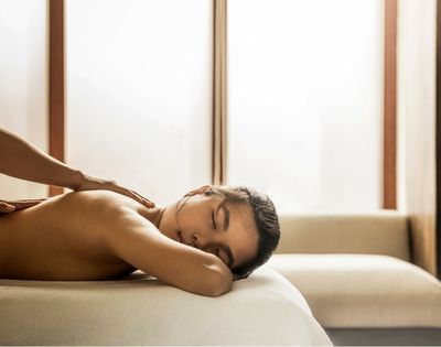 Shirtless man rests face down on a sofa while receiving a back massage in a softly lit room, creating a calm, relaxing atmosphere.