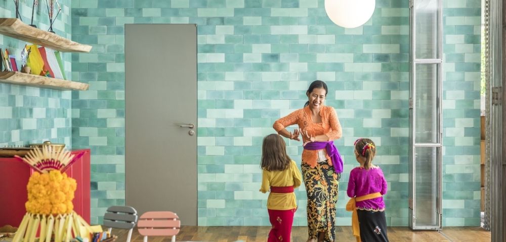Woman in a colorful kitchen helps two children adjust traditional outfits, creating a warm, joyful family moment amid turquoise tiles and crafts.
