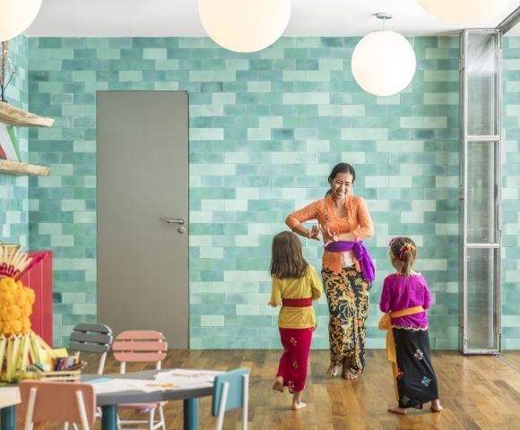 Woman in a colorful kitchen helps two children adjust traditional outfits, creating a warm, joyful family moment amid turquoise tiles and crafts.