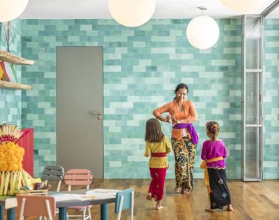 Woman in a colorful kitchen helps two children adjust traditional outfits, creating a warm, joyful family moment amid turquoise tiles and crafts.