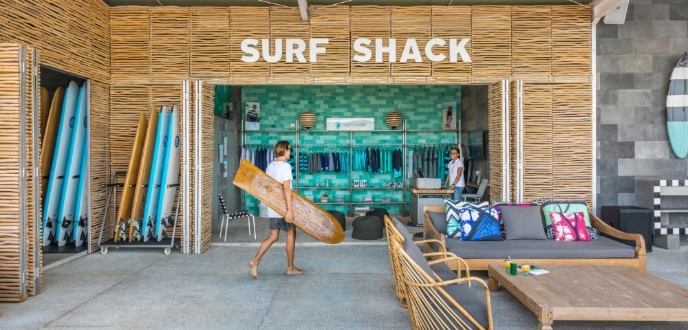 Bright beach resort surf shack with bamboo walls, colorful surfboards, beach gear, lounge chairs, and a person carrying a board.