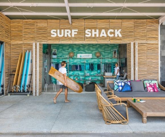 Bright beach resort surf shack with bamboo walls, colorful surfboards, beach gear, lounge chairs, and a person carrying a board.