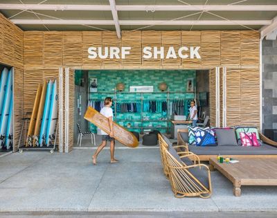 Bright beach resort surf shack with bamboo walls, colorful surfboards, beach gear, lounge chairs, and a person carrying a board.