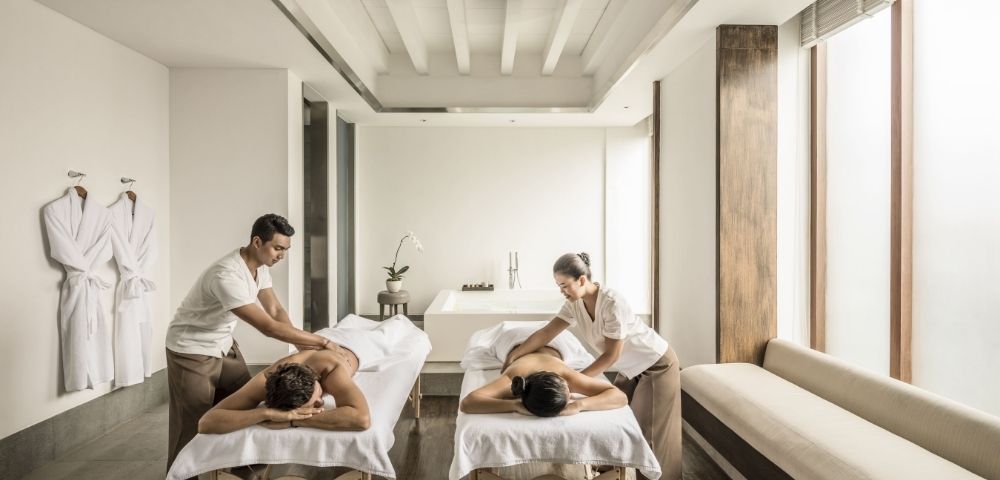 Bright, serene spa room with two massage tables; therapists treat clients as white robes hang nearby and sunlight creates a calming mood.