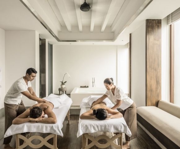 Bright minimalist spa with two massage therapists treating clients on white tables; robes hang on the wall, conveying a calm, serene mood.