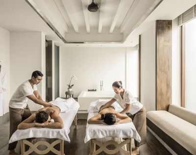 Bright minimalist spa with two massage therapists treating clients on white tables; robes hang on the wall, conveying a calm, serene mood.