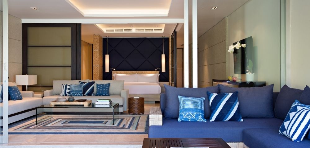 Modern luxury hotel suite with a blue sectional sofa, patterned pillows, glass coffee table, and bedroom beyond; calm, elegant interior.