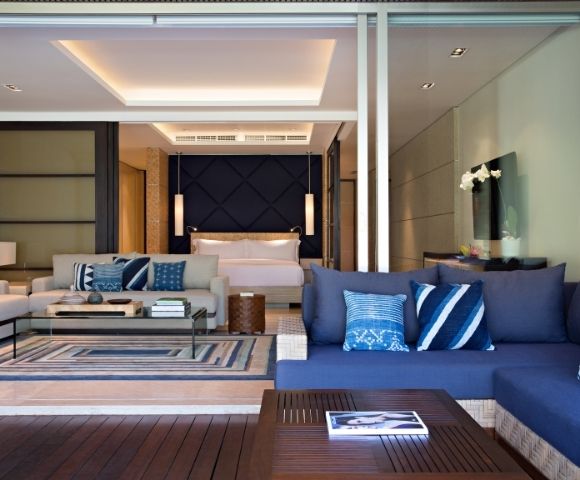 Modern luxury hotel suite with a blue sectional sofa, patterned pillows, glass coffee table, and bedroom beyond; calm, elegant interior.