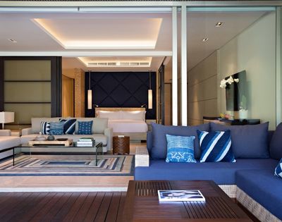 Modern luxury hotel suite with a blue sectional sofa, patterned pillows, glass coffee table, and bedroom beyond; calm, elegant interior.