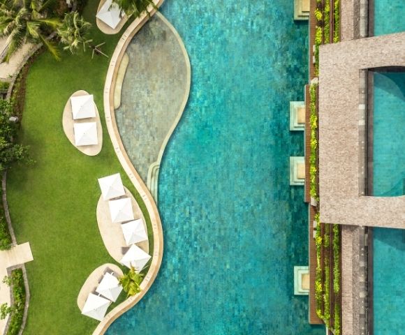 Aerial view of a luxury resort with a turquoise pool, curved swim area, white loungers, manicured lawns, palms, and modern architecture.