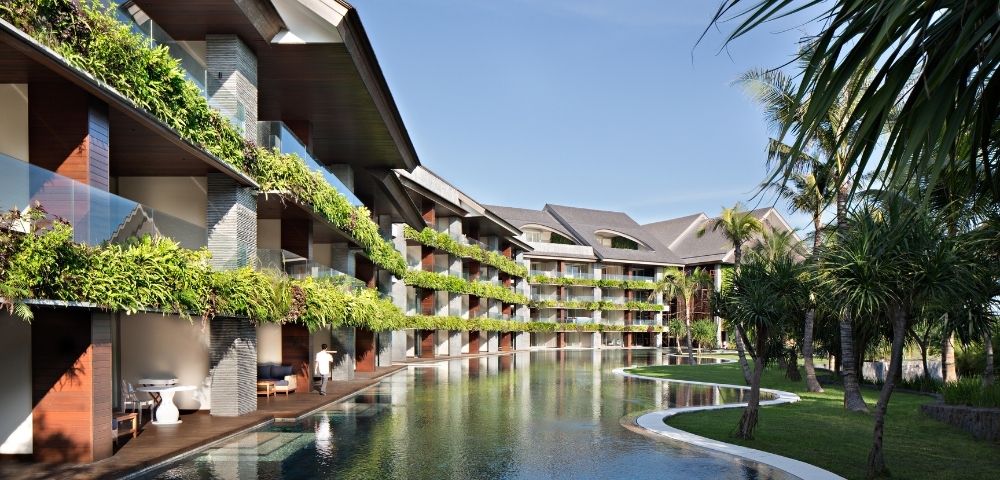 Modern tropical resort with lush balconies, calm reflecting pool, palm trees, and curving paths under a clear blue sky, creating a serene mood.