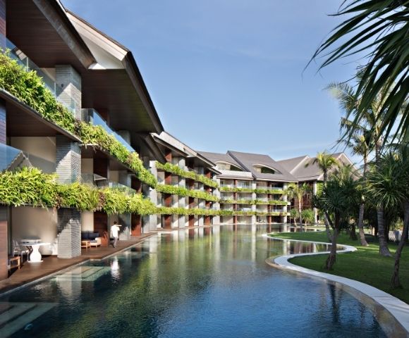 Modern tropical resort with lush balconies, calm reflecting pool, palm trees, and curving paths under a clear blue sky, creating a serene mood.