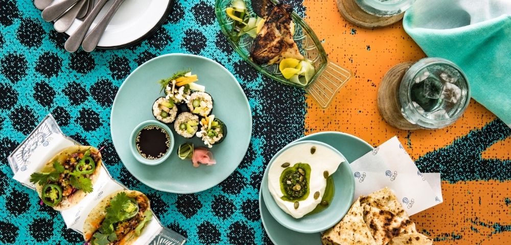 Overhead spread of sushi, avocado toast, dessert, drinks, and cutlery on a turquoise-and-orange patterned tablecloth; lively dining scene.